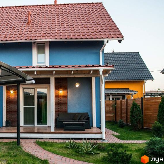 Будинок блакитного кольору з червоною черепицею та білою терасою, освітленою ліхтарями. Навколо будинку зелений газон з доріжкою з бруківки.