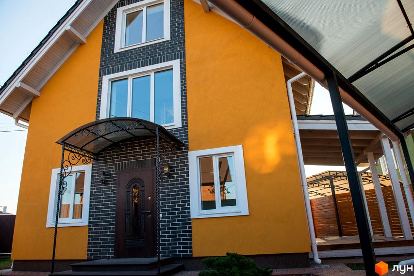 Фасад будинку Котеджного містечка Osocor Scandi House з жовтою штукатуркою та темно-сірою цеглою, білі вікна. Поруч вимощена доріжка та навіс.