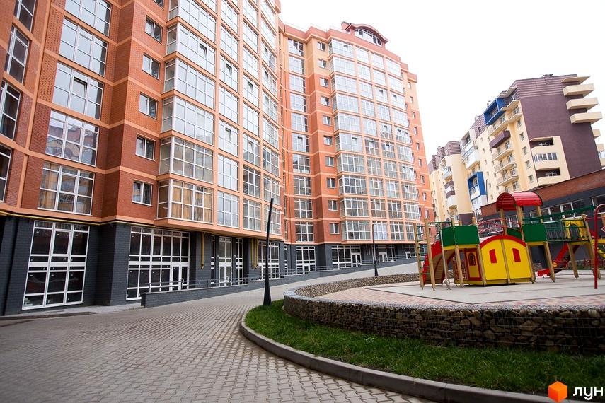 Багатоповерховий житловий будинок ЖБК Перспектива з цегляно-помаранчевим фасадом та великими вікнами. Перед будинком облаштована територія з дитячим майданчиком та бруківкою.