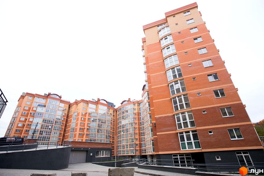 Багатоповерхові будинки ЖБК Перспектива з цегляними фасадами помаранчевих та коричневих відтінків, великими вікнами. Прибудинкова територія включає доріжки та в'їзд до паркінгу.