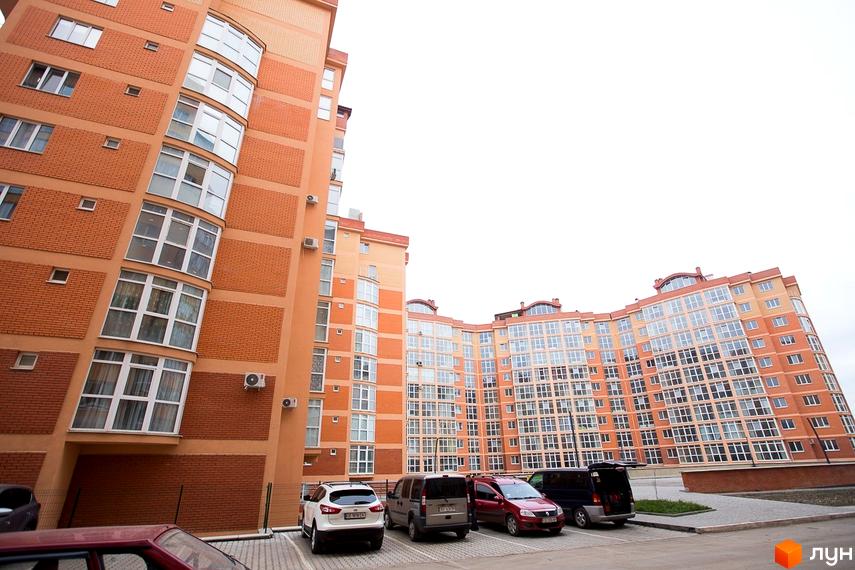 Житловий комплекс Перспектива з вигнутими помаранчевими фасадами та численними вікнами. Перед будинком розташований асфальтований паркінг з автомобілями.