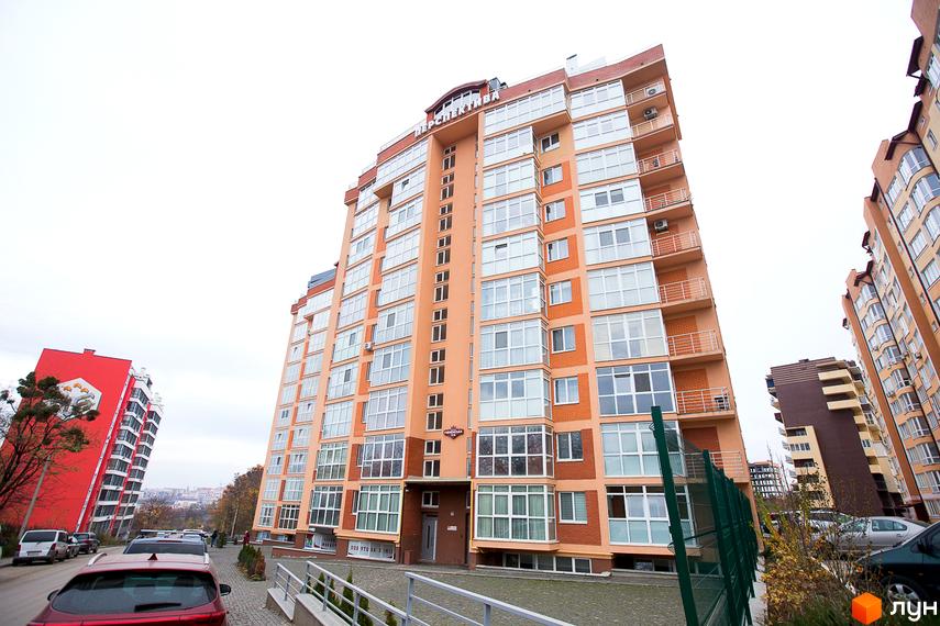 Багатоповерховий житловий комплекс Перспектива світло-помаранчевого кольору з цегляними вставками та балконами. Прибудинкова територія має парковку.