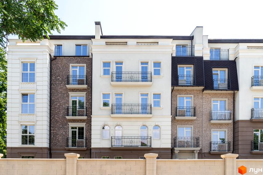 Фасад житлового комплексу CHELSEA Club House з білою та цегляною обробкою, балконами та великими вікнами. Будівля розташована на тлі блакитного неба, частково прихована зеленню дерев. Перед комплексом видно кам'яний паркан.