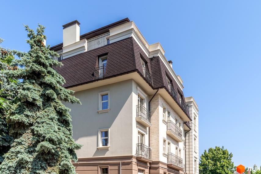 Фасад житлового комплексу CHELSEA Club House з мансардним дахом та балконами. Навколо будівлі зелені дерева та блакитне небо.