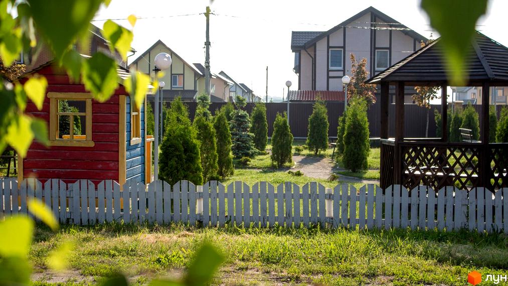 Будинок з червоно-синім фасадом та білим парканом на тлі котеджного містечка Вишневий хуторок. На території газони, дерева та альтанка.