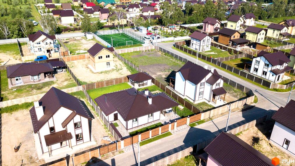 Зображення житлового комплексу КМ Вишневий хуторок. На фото видно котеджі з коричневими дахами та білими стінами, огороджені дерев'яними парканами. На території є газони та доріжки.