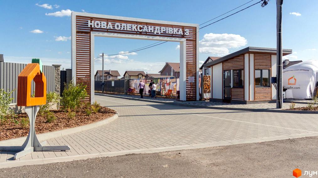 Вхід до Котеджного містечка Нова Олександрівка 3 з великою аркою та офісом продажу. На території бруківка, доглянуті зелені насадження та будинки на задньому плані.