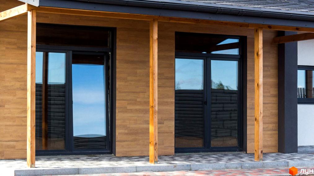 Фасад будинку Котеджного містечка Нова Олександрівка оздоблений деревом, з великими чорними вікнами та дерев'яними опорами. Прибудинкова територія вимощена плиткою.