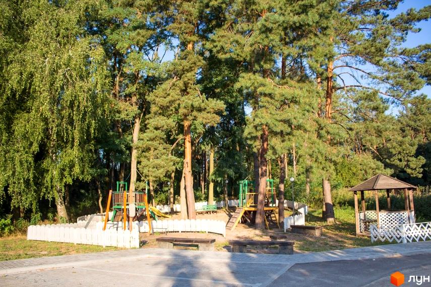 Зображення дитячого майданчика в Містечку котеджів Лісове озеро. На території комплексу багато дерев та є альтанка.