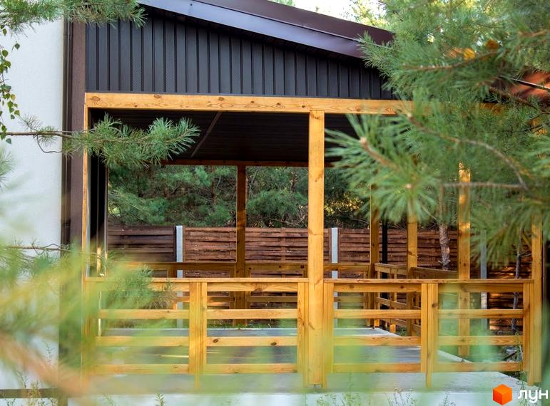 Котеджне містечко Лісове озеро. Зображення дерев'яної альтанки з поручнями та дахом. На задньому плані видно зелень дерев.