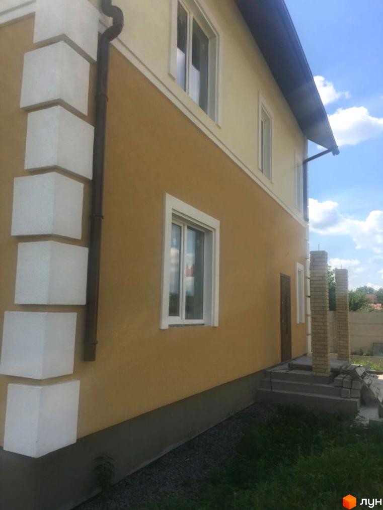 Фасад будинку Котеджного містечка Шевченківський виконаний у світло-коричневих тонах з білими декоративними елементами. Навколо будинку зелена трава та доріжка з гравію.