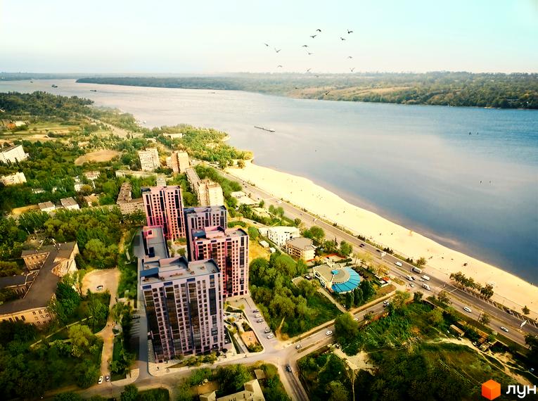 Житловий комплекс Legenda: високі будівлі рожево-фіолетових відтінків з багатьма вікнами, розташовані біля широкої річки з пляжем та зеленими насадженнями.