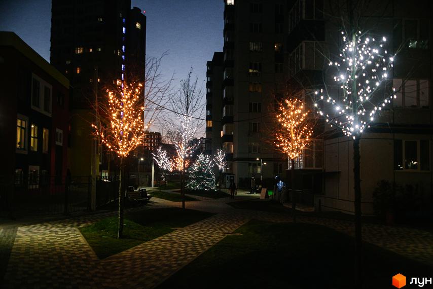 Житловий комплекс Синергія-3: багатоповерхові будинки ввечері, а також прибудинкова територія з деревами, прикрашеними гірляндами.