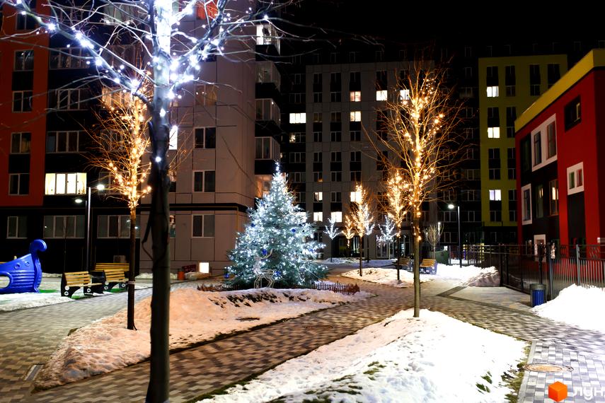 Житловий комплекс Синергія-3 взимку. Багатоповерхові будинки з яскравими фасадами та святково освітлена прибудинкова територія з ялинкою та деревами.