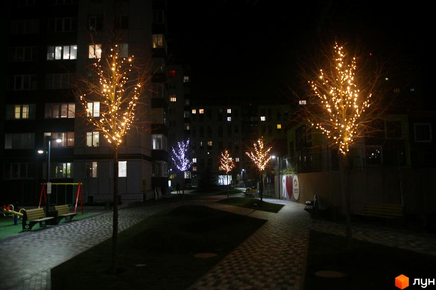 Житловий комплекс Синергія-3 вночі з освітленими вікнами. Благоустрій території включає дерева з гірляндами, лавки та дитячий майданчик.