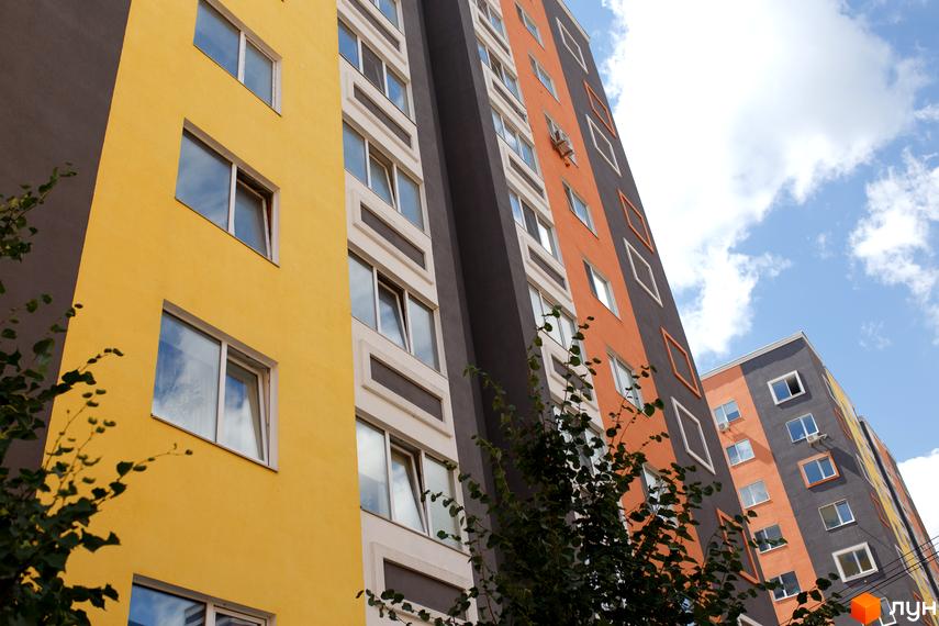 Житловий комплекс Синергія-2 з яскравим фасадом та великими вікнами. На фото зображено частину будинку на фоні блакитного неба з хмарами.