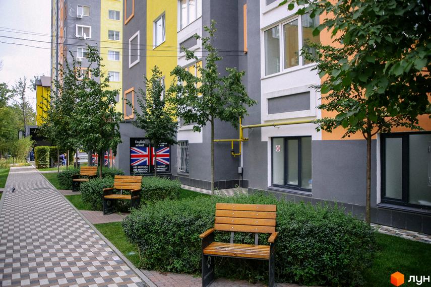 Зовнішній вигляд будівлі житлового комплексу Синергія-2 з фасадом у сірих та жовтих тонах. На території розташовані лавки для відпочинку та зелені насадження.