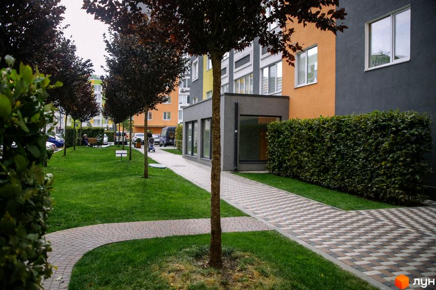 Житловий комплекс Синергія-2 з фасадом в сіро-жовтих тонах. Доглянута прибудинкова територія з газоном, деревами та тротуарами.