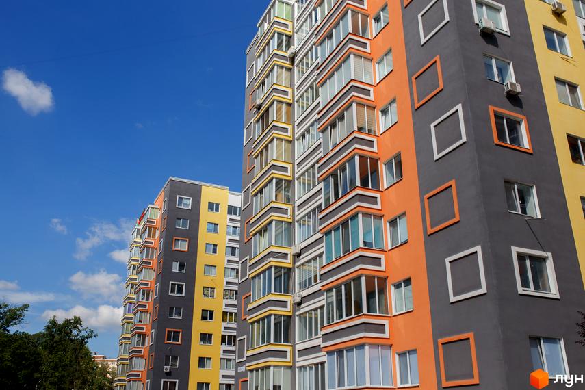 Яскравий багатоповерховий будинок ЖК Синергія-2 з великими вікнами та балконами на тлі блакитного неба. Фасад будівлі пофарбовано в різні кольори: помаранчевий, сірий та жовтий.