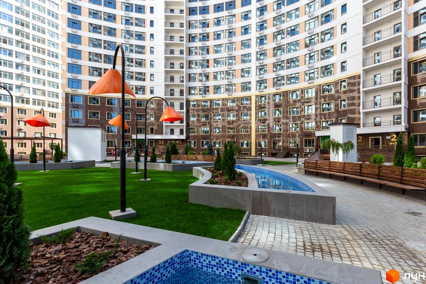 Фото внутрішнього двору Новобудови Тридцять друга перлина. На фото будівля з великою кількістю вікон та зелена прибудинкова територія з лавками та басейном.