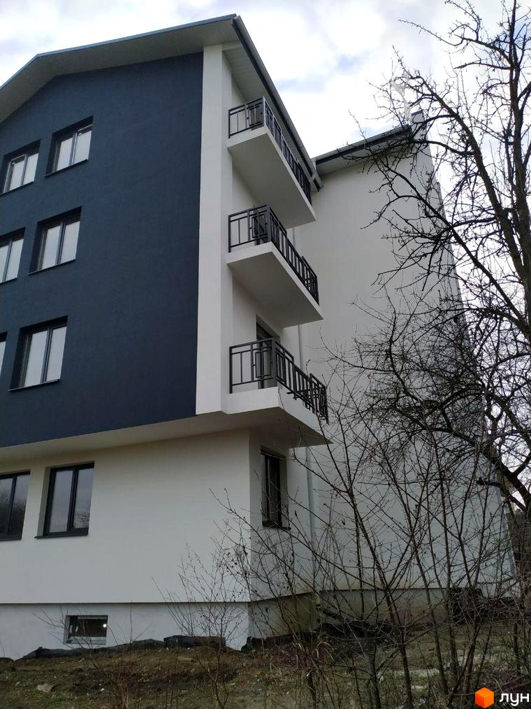 Житловий комплекс Панський острів з темно-сірим та білим фасадом, балконами з чорними перилами. Навколо будинку видно голі дерева та ґрунт.