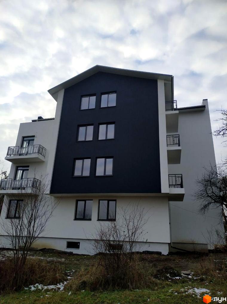 Зображення фасаду Житлового комплексу Панський острів з білими та темно-сірими секціями, балконами та вікнами. На передньому плані видно суху траву та кущі, а також ділянки зі снігом.