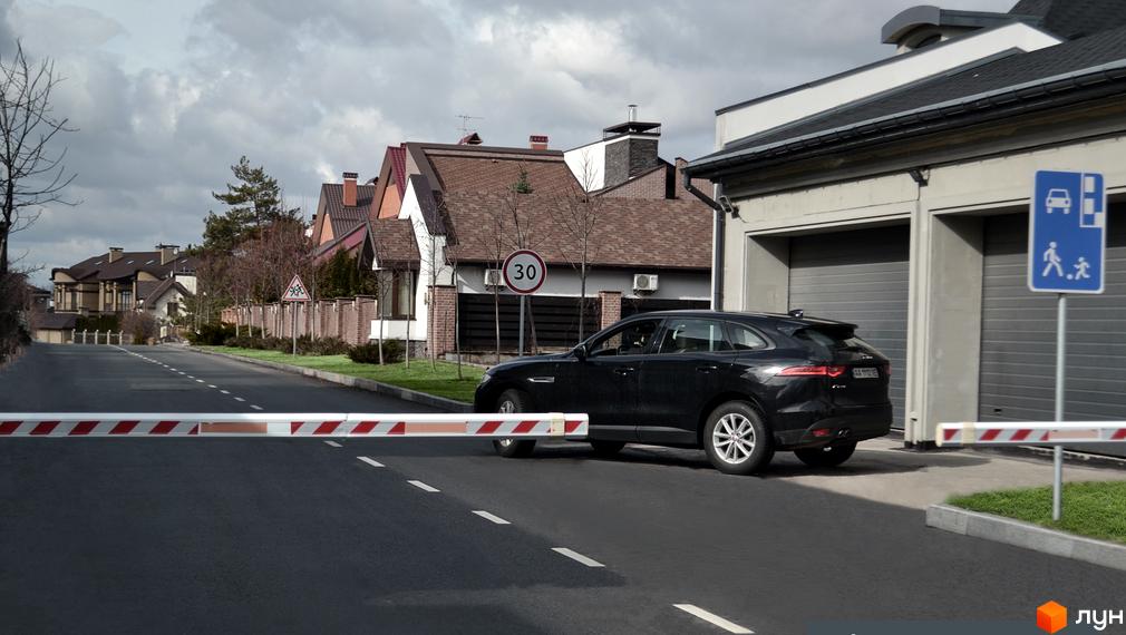 Зображення сторінки житлового комплексу КМ Lisnyky Club. На фото видно чорний автомобіль біля гаражів, а також дорогу з шлагбаумом та будинки з коричневими дахами.