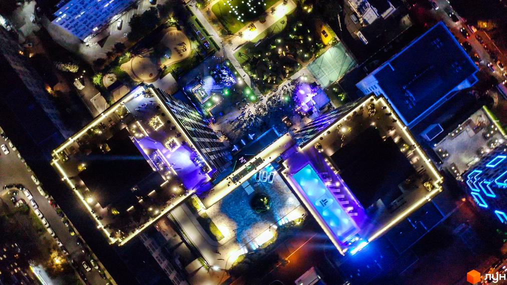 Нічний вигляд житлового комплексу Tetris Hall з висоти пташиного польоту. Освітлені будівлі та басейн на даху, зелена зона та спортивні майданчики.