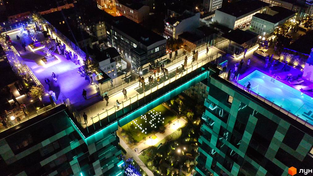 Житловий комплекс Tetris Hall у Києві. Нічний вигляд на будівлі з басейном на даху та підсвіченим мостом між будинками. Зелена територія з освітленням.