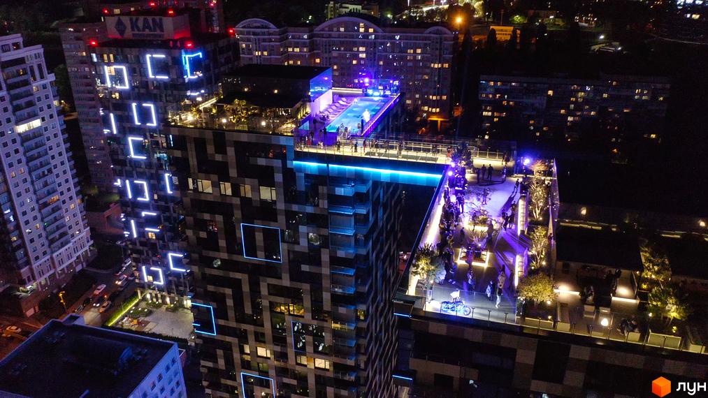 Житловий комплекс Tetris Hall з яскравою підсвіткою фасаду та басейном на даху. Навколо комплексу розташовані зони відпочинку з деревами та людьми.