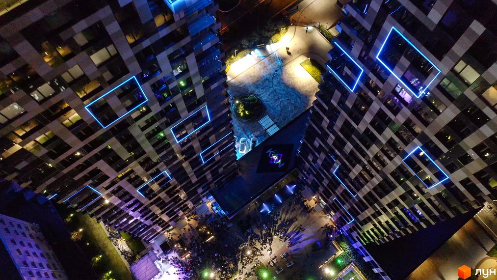Житловий комплекс Tetris Hall з яскравою синьою підсвіткою вікон. Навколо комплексу розташовані зони відпочинку з людьми та освітленням.