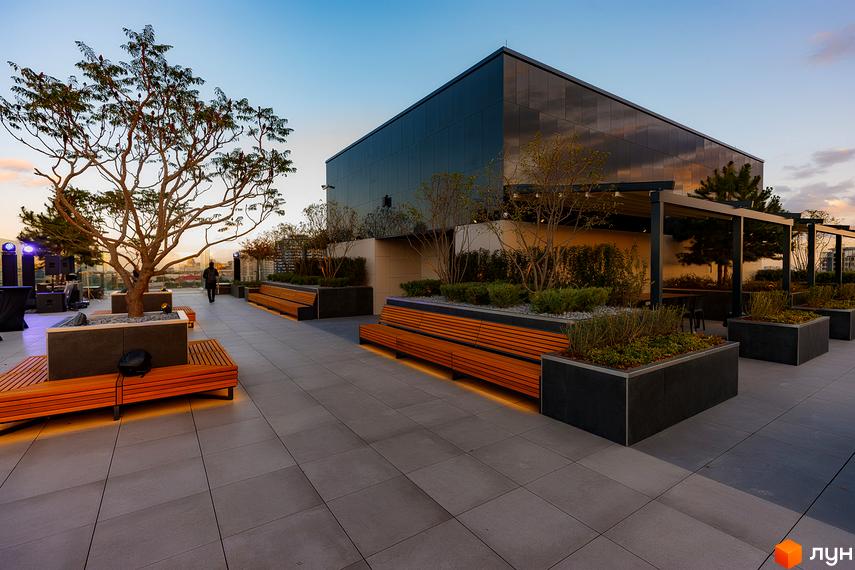 Сучасна тераса житлового комплексу Tetris Hall з дерев'яними лавами та озелененням. Вид на будівлю з дзеркальним фасадом на заході сонця.