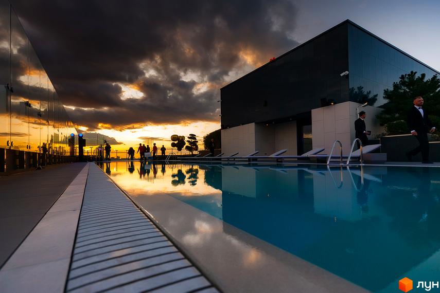 Житловий комплекс Tetris Hall з басейном на даху та людьми на фоні заходу сонця. Сучасна архітектура новобудови з панорамним склінням.