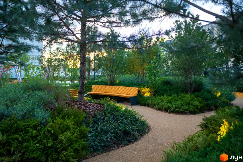 Житловий комплекс Tetris Hall: зелена зона з лавкою та доріжкою. Територія навколо будинку з деревами та кущами.