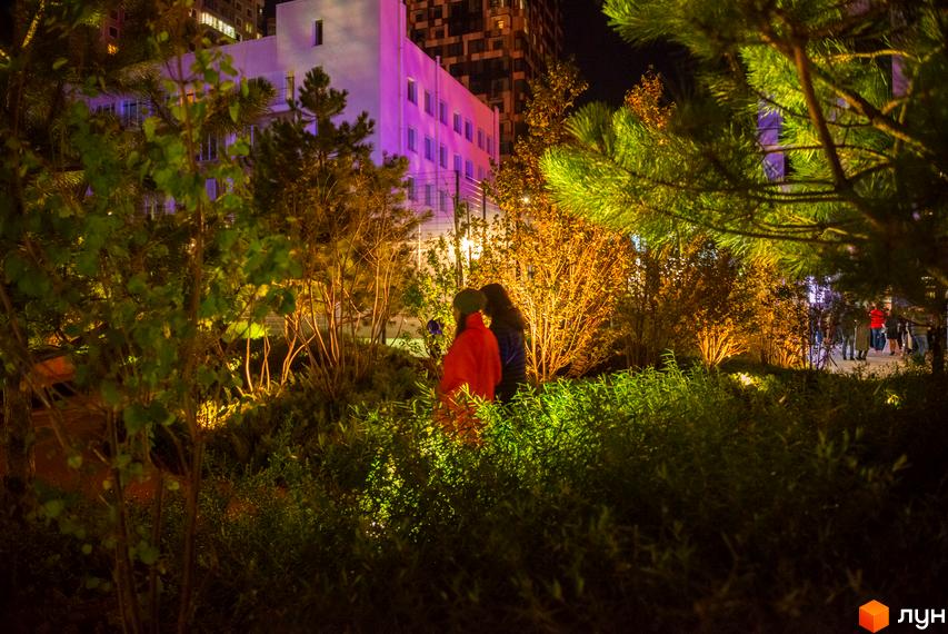 Житловий комплекс Tetris Hall вночі. Освітлена будівля з фіолетовим підсвічуванням та зелена прибудинкова територія з деревами та кущами.