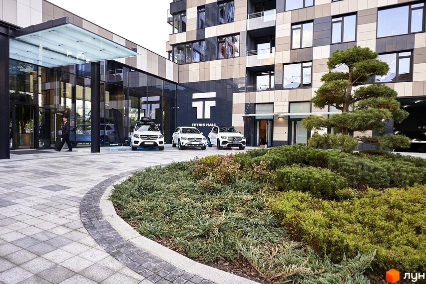 Житловий комплекс Tetris Hall з фасадом з панелей та скляними елементами. Перед будівлею розташовані три білих автомобілі Mercedes-Benz та доглянутий зелений ландшафт з кущів та дерев.