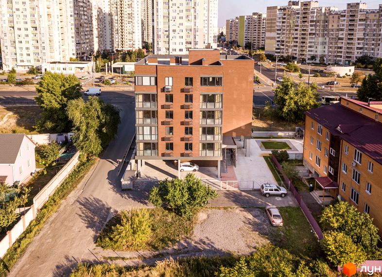 Клубний будинок „Деснянський DeLuxe“ з цегляним фасадом та великими вікнами. Навколо будинку розташована прибудинкова територія з зеленими насадженнями та парковкою.