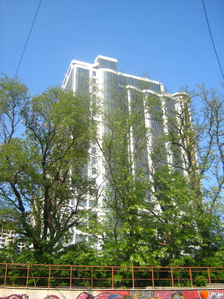 Високий білий будинок Житлового комплексу Гагарін Плаза 1 з великими вікнами. Перед ним розташовані густі зелені дерева та металева огорожа.