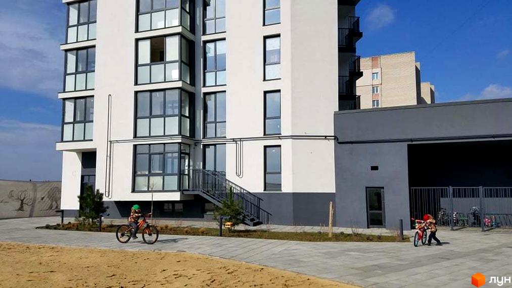 Білий багатоповерховий будинок з темними вікнами та сходами. Прибудинкова територія Житлового комплексу Басів схил з доріжками та дітьми на велосипедах.