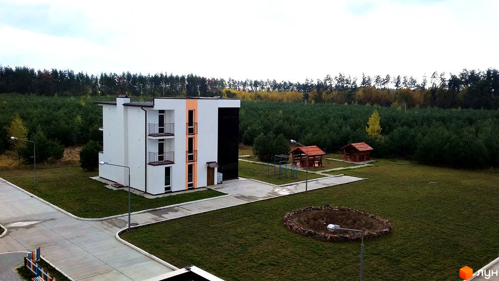 Зображення триповерхового будинку в Житловому комплексі Ясний. На території є зелений газон, дерева та дві альтанки.