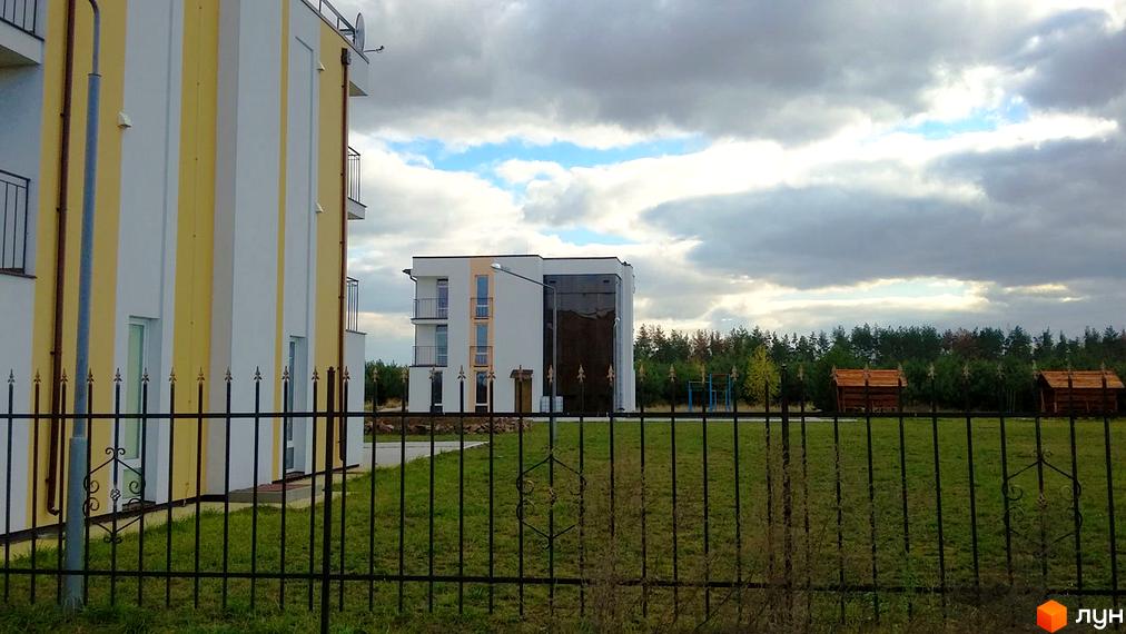 Зображення фасаду будинку в Житловому комплексі Ясний. На передньому плані металева огорожа, за нею газон та дерева.