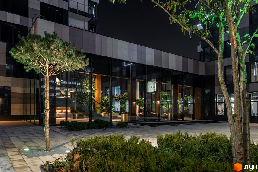 Житловий комплекс Tetris Hall. Нічна фотографія фасаду будівлі з великими вікнами та підсвіченими деревами. Територія навколо комплексу викладена плиткою, з доглянутими кущами та деревами.