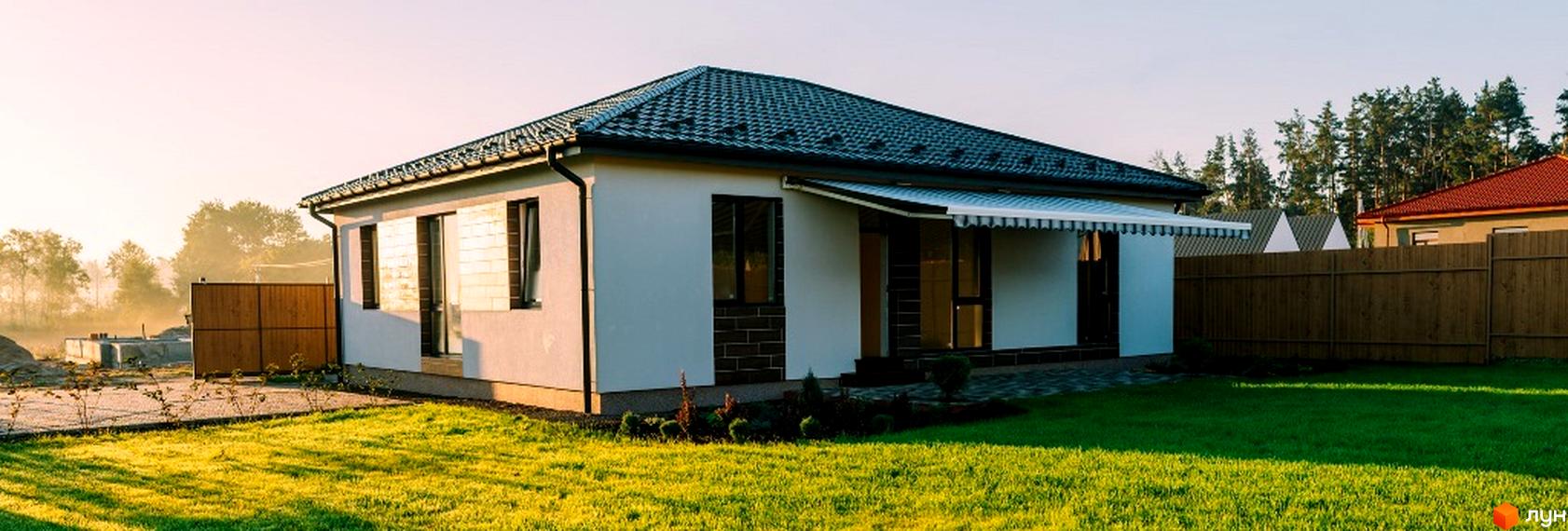 Котеджне містечко КМ Villa Grace: одноповерховий будинок з темним дахом та білим фасадом, облаштований навісом, на тлі зеленого газону та дерев.