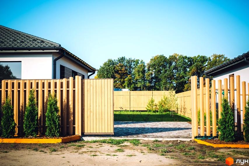Котеджне містечко КМ Villa Grace: білий будинок з дерев'яним парканом та зеленими туями. Прибудинкова територія з бруківкою та деревами.