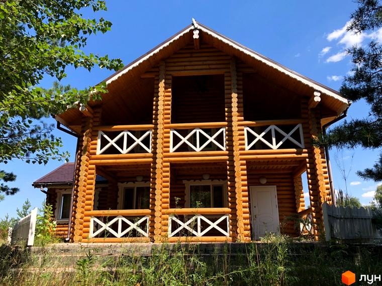 Двоповерховий дерев'яний будинок з балконами та білими перилами на тлі блакитного неба. Оточений зеленню та травою.