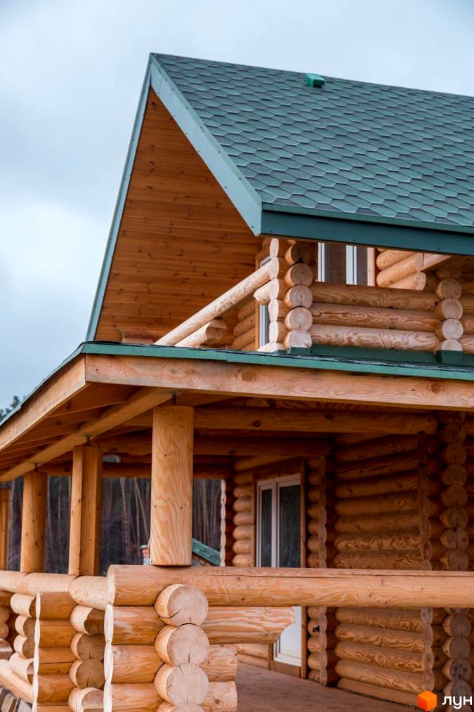 Котеджне містечко Святобор: дерев'яний будинок з брусу з зеленою черепичною покрівлею та верандою. Навколо будинку видно ліс та небо.