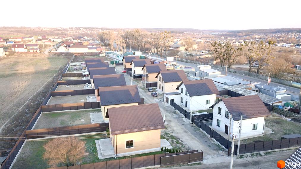 Котеджне містечко КМ Green House: акуратні двоповерхові будинки з коричневими дахами та доглянутими ділянками, огородженими парканами. Навколо зелені насадження та дороги.