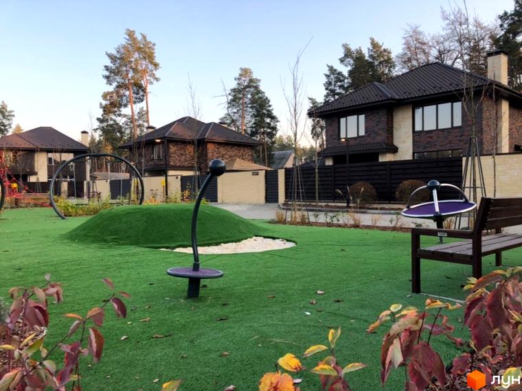 Котеджне містечко Zahrava Village: сучасний дитячий майданчик з штучним покриттям та будинок на фоні. Зелена зона відпочинку з лавкою та ігровими елементами.