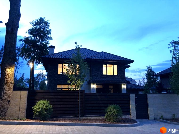 Котеджне містечко Zahrava Village. Фасад будинку з темної цегли підсвічений ліхтарями, навколо огороджена територія з деревами та кущами.