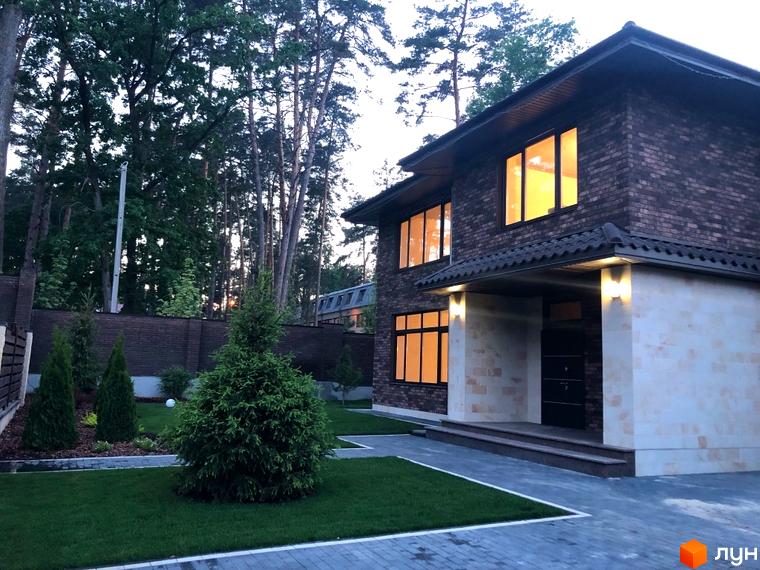 Котеджне містечко КМ Zahrava Village. Двоповерховий будинок з коричневої цегли з великими вікнами, що виходять на доглянутий двір з газоном та ялинками.